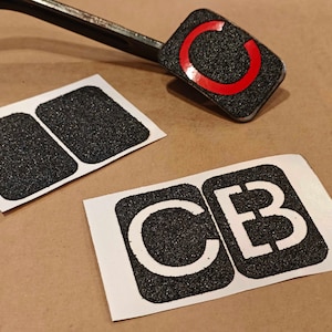 Peut inclure: Trois autocollants adhésifs en bande de roulement noire et blanche. Un autocollant a un cercle rouge avec un contour blanc. Les deux autres autocollants ont les lettres "C" et "B" en blanc.