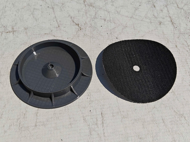May include: Two-piece set featuring a dark gray, circular base with a central cone and a black, circular pad with a central hole. The items are likely components of a tool or device.