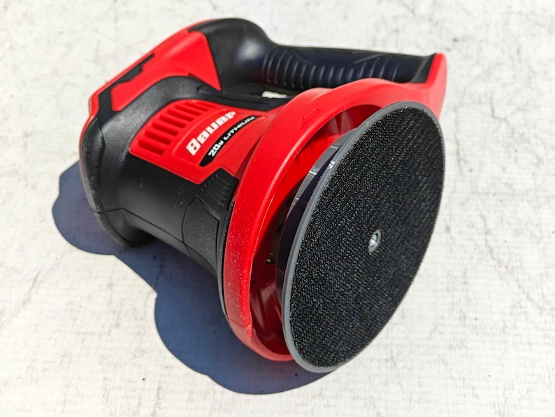 May include: A red and black Bauer 20V Lithium random orbital sander. The sander has a black grip, a red housing, and a round, black sanding pad. The Bauer logo is visible on the side.