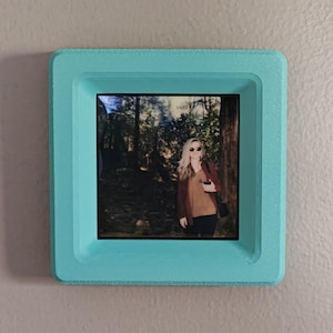 May include: A turquoise picture frame with a photo of a person wearing sunglasses and a brown sweater standing in a forest.