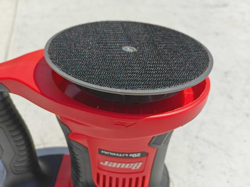 May include: A red and black Bauer 20V Lithium sander. The sander has a round, black sanding pad with a hook and loop surface. The body of the sander is red with black accents and the Bauer logo.