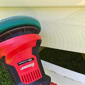 May include: A red and black Bauer 20V Lithium polisher with a green buffing pad. The tool is designed for automotive detailing and surface restoration. The image shows the polisher in use, with a section of a light-colored surface visible in the background.