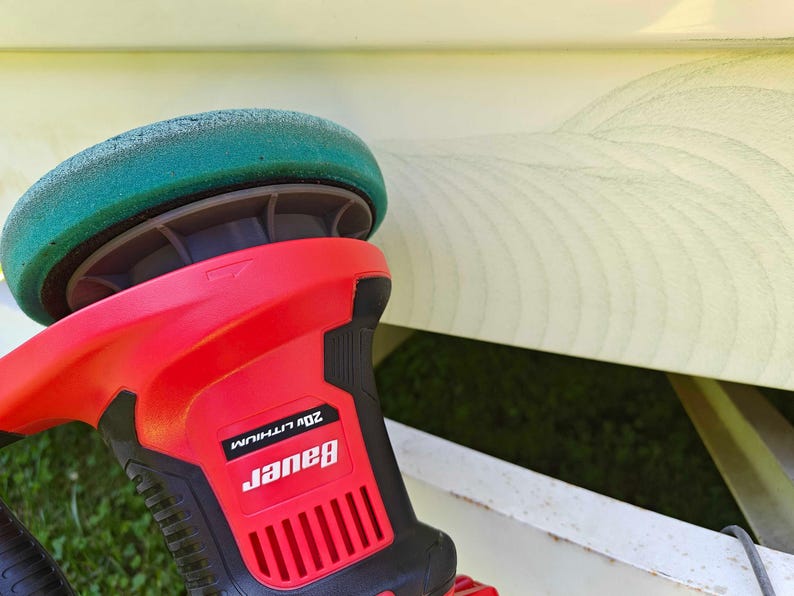 May include: A red and black Bauer 20V Lithium polisher with a green buffing pad. The tool is designed for automotive detailing and surface restoration. The image shows the polisher in use, with a section of a light-colored surface visible in the background.