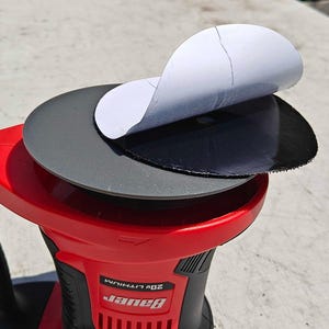 May include: A red and black power sander with a round sanding pad. A black sanding disc is partially peeled back, revealing a white backing. The sander has the brand name "Bauer" printed on it.