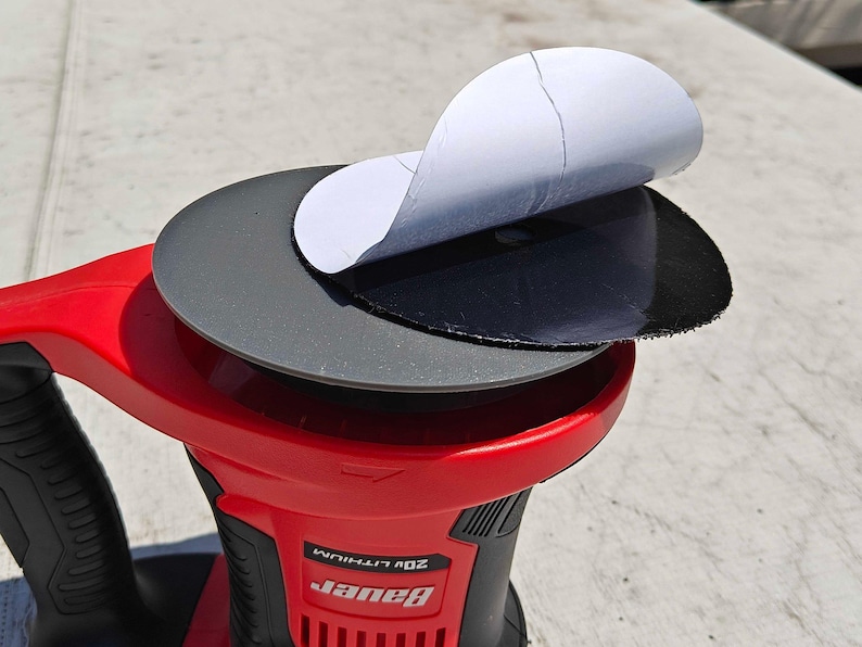 May include: A red and black power sander with a round sanding pad. A black sanding disc is partially peeled back, revealing a white backing. The sander has the brand name "Bauer" printed on it.