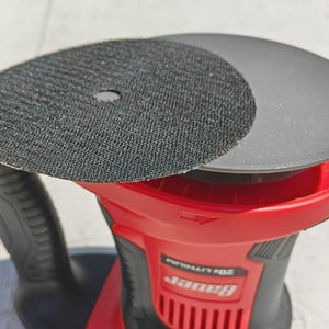 May include: A red and black power sander with two circular sanding pads. One pad is black with a textured surface, and the other is gray. The sander has a black handle and the brand name "Bauer" is visible.