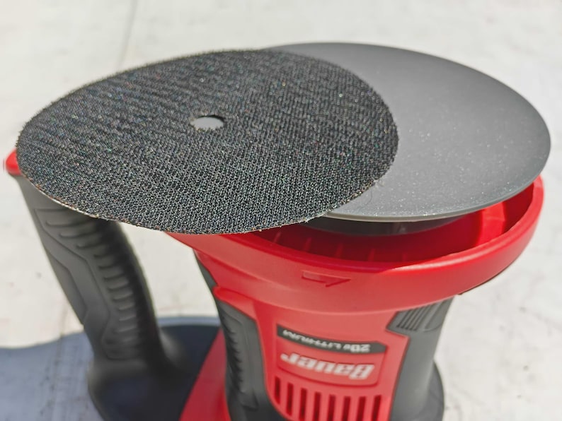 May include: A red and black power sander with two circular sanding pads. One pad is black with a textured surface, and the other is gray. The sander has a black handle and the brand name "Bauer" is visible.