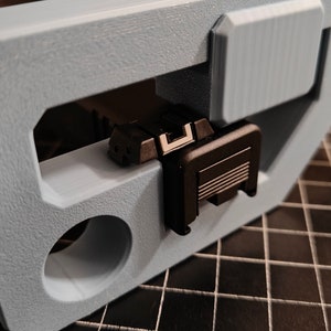 May include: A black plastic part with a textured surface and a small rectangular opening. The part is held in a light blue plastic clamp.