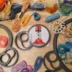 May include: A close-up of a cross-stitch project of a red and white lighthouse on a white background. The project is in a round embroidery hoop. There are various colors of embroidery floss in the background.