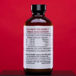 May include: A brown glass bottle with a black cap, labeled "VULCAN'S TREASURE ORMUS CONCENTRATE." The label lists mineral content per teaspoon, including iron, magnesium, and sodium. The bottle is against a red background.