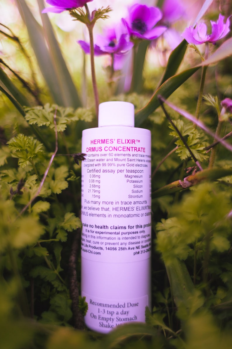 Puede incluir: Una botella blanca de "HERMES' ELIXIR ORMUS CONCENTRATE" rodeada de follaje verde y flores moradas. La etiqueta incluye texto sobre los ingredientes y la dosis recomendada. La botella mide unos 15 cm de altura.