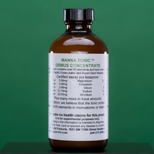 May include: A brown glass bottle with a black cap and a white label. The label reads "MANNA TONIC ORMUS CONCENTRATE" and lists ingredients and assay information. The bottle is filled with a clear liquid.