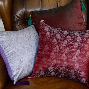 Puede incluir: Tres cojines decorativos con patrones intrincados y borlas. Uno es de color lavanda claro con un diseño floral, otro es de color burdeos oscuro con un patrón floral, y el tercero es de color rojo oscuro con un patrón floral. Los cojines están en un sillón de cuero marrón.
