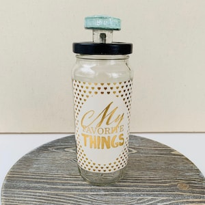 May include: A clear glass jar with a black lid and a light blue glass knob. The jar has a white label with gold hearts and the text "My Favorite Things".