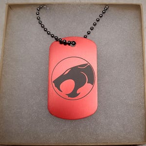 May include: A red dog tag pendant with a black chain. The tag features a black graphic of a stylized cat's head inside a circle. The pendant is rectangular with rounded corners and is presented in a cardboard box.