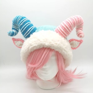 May include: A blue and pink crocheted hat with white fur trim and curled horns. The hat is on a white mannequin head with pink hair.