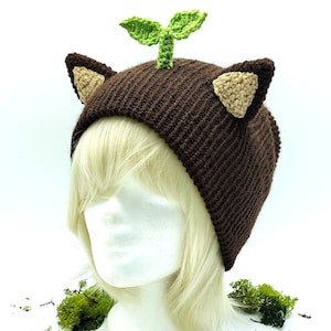 Könnte beinhalten: Ein brauner gehäkelter Beanie mit beigefarbenen Katzenohren und einem grünen gehäkelten Spross oben. Der Hut wird auf einem weißen Schaufensterkopf mit blonden Haaren präsentiert, umgeben von grünem Moos.