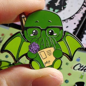 Op de afbeelding: Groene emaille pin met een cartoon Cthulhu-personage met een bril, dobbelstenen en een notitieblok. De pin heeft vleugels en staat tegen een groen-roze achtergrond. Andere pins zijn zichtbaar.