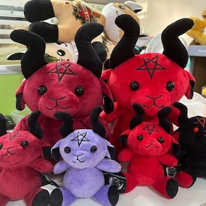 Könnte beinhalten: Fünf Plüschtiere in Form von roten und lila Ziegen mit schwarzen Hörnern und einem Pentagramm auf der Stirn. Die Ziegen sitzen auf einer weißen Oberfläche.