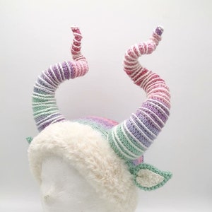 May include: A whimsical, handmade hat featuring large, spiraling horns in shades of pink, purple, and green. The hat has a fluffy, cream-colored brim and small, green crocheted ears. The horns are made of yarn.