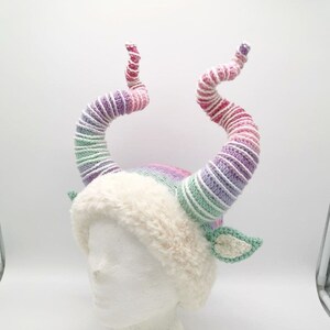May include: A whimsical, handmade hat with large, spiraling horns in shades of pink, purple, and green. The hat features a fluffy, cream-colored brim and small, green crocheted leaf accents, creating a fantasy-themed accessory.