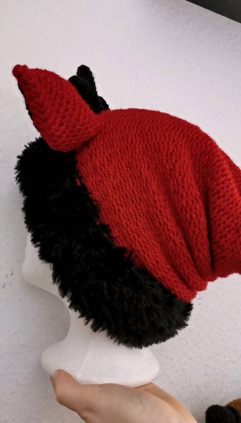 Puede incluir: Un gorro de punto rojo y negro con un ribete negro y peludo. El gorro tiene un dise&ntilde;o puntiagudo en forma de oreja en la parte superior. El gorro se exhibe en una cabeza de maniqu&iacute; blanca. El gorro est&aacute; hecho de un material suave y texturizado.