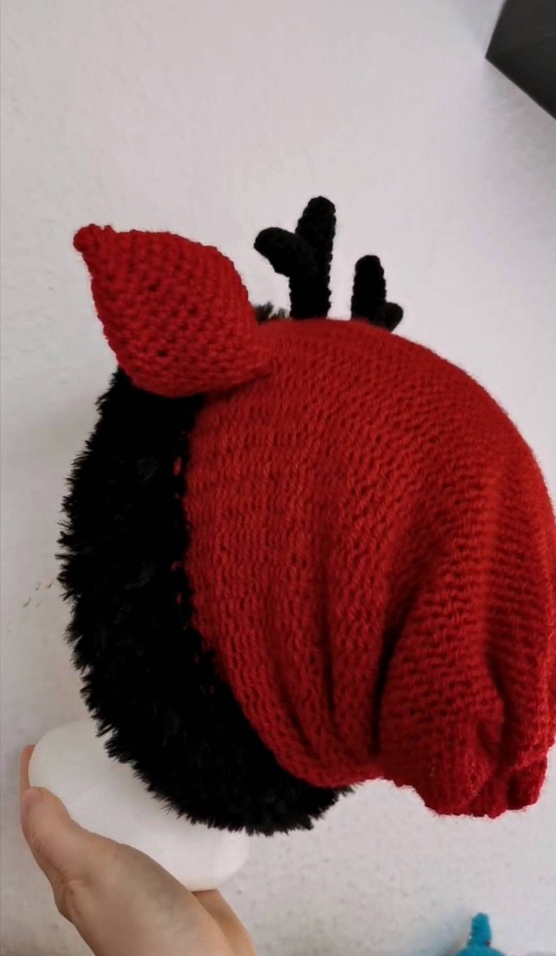 Puede incluir: Un gorro de punto rojo y negro hecho a mano con un dise&ntilde;o &uacute;nico. El gorro presenta una parte superior de punto rojo con piel sint&eacute;tica negra y peque&ntilde;os cuernos negros. El gorro est&aacute; dise&ntilde;ado para ser usado en la cabeza.