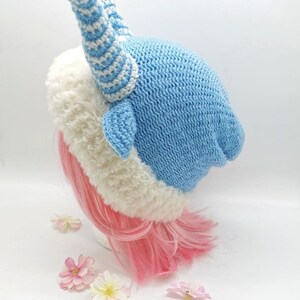 May include: A blue and white crocheted hat with a white fur trim and a large, striped horn on top. The hat is on a pink wig.
