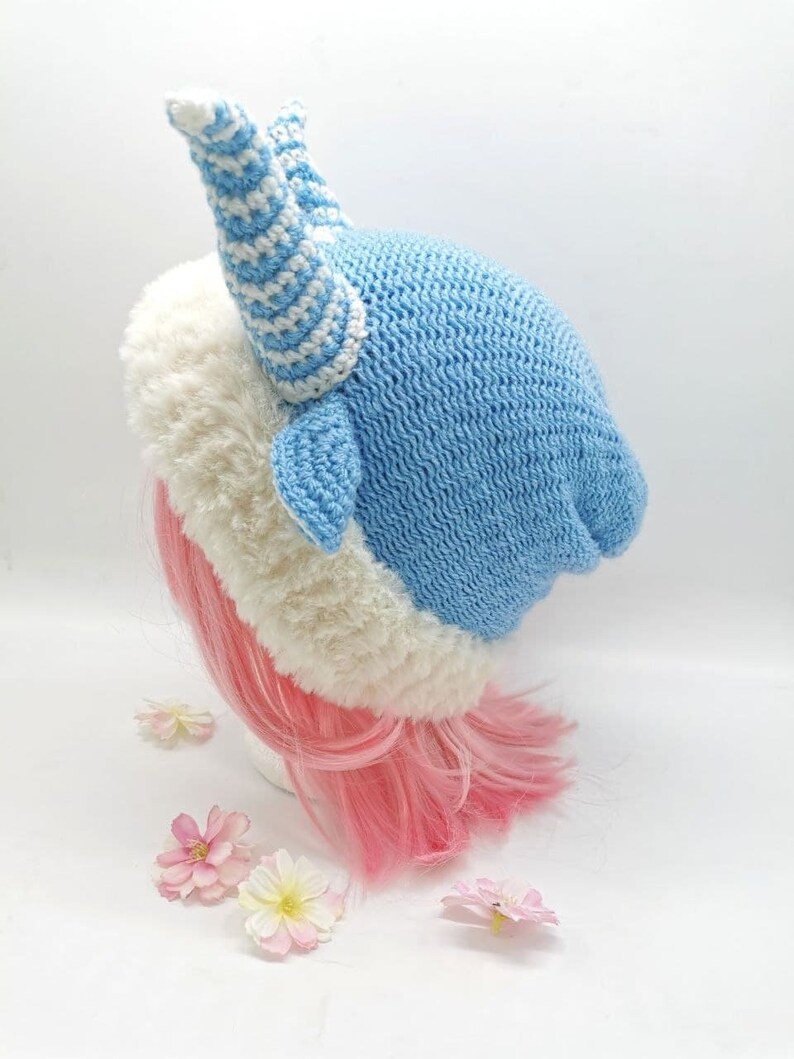 May include: A blue and white crocheted hat with a white fur trim and a large, striped horn on top. The hat is on a pink wig.