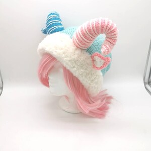 K&ouml;nnte beinhalten: Eine gestrickte M&uuml;tze mit blauen und rosa Streifen, wei&szlig;em Pelzbesatz und zwei gekr&auml;uselten H&ouml;rnern. Die M&uuml;tze wird von einem Mannequin mit rosa Haaren getragen.