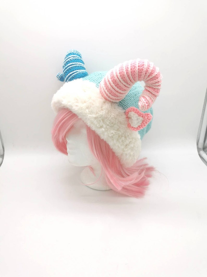 K&ouml;nnte beinhalten: Eine gestrickte M&uuml;tze mit blauen und rosa Streifen, wei&szlig;em Pelzbesatz und zwei gekr&auml;uselten H&ouml;rnern. Die M&uuml;tze wird von einem Mannequin mit rosa Haaren getragen.