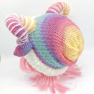 Puede incluir: Un gorro de punto de arco&iacute;ris con un borde de piel sint&eacute;tica blanca y dos cuernos rizados. Los cuernos est&aacute;n hechos de hilo blanco y rosa.