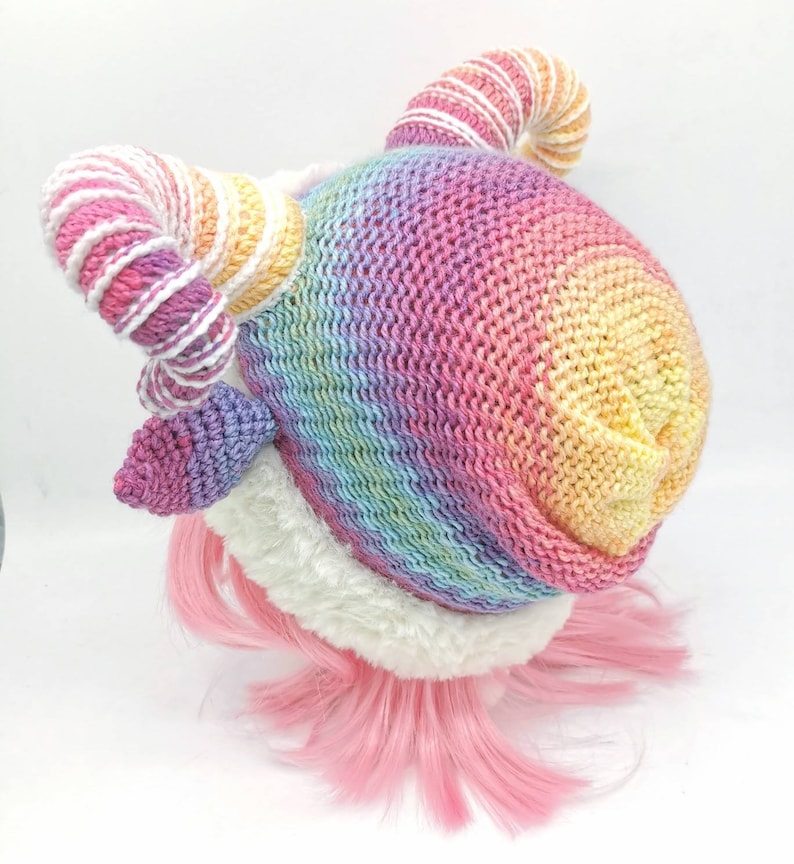 Puede incluir: Un gorro de punto de arco&iacute;ris con un borde de piel sint&eacute;tica blanca y dos cuernos rizados. Los cuernos est&aacute;n hechos de hilo blanco y rosa.