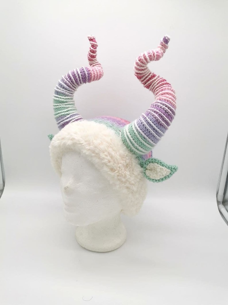 May include: A whimsical, handmade hat featuring large, spiraling horns in shades of pink, purple, and green. The hat has a fluffy, cream-colored brim and small, green crocheted ears. The horns are made of yarn.