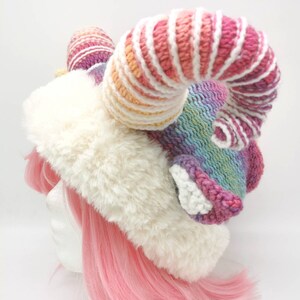 Puede incluir: Un gorro de piel sint&eacute;tica blanca con un cuerno de unicornio de crochet arco&iacute;ris. El cuerno est&aacute; rayado con hilo rosa, blanco y amarillo.