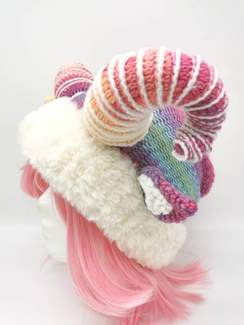 Puede incluir: Un gorro de piel sint&eacute;tica blanca con un cuerno de unicornio de crochet arco&iacute;ris. El cuerno est&aacute; rayado con hilo rosa, blanco y amarillo.