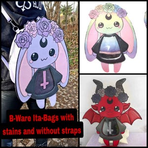 Puede incluir: Tres ita-bags con diferentes diseños. La primera es una conejita morada con una cruz negra y un halo rosa. La segunda es una conejita arcoíris con una cruz negra y un halo arcoíris. La tercera es un diablo rojo con una cruz negra y un halo negro. Las tres bolsas tienen una ventana transparente en la parte delantera. B-Ware Ita-Bags con manchas y sin correas.