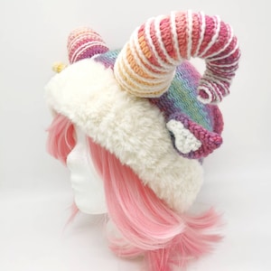 Puede incluir: Un gorro de crochet rosa y blanco con un cuerno grande, rizado y de colores arco&iacute;ris. El gorro est&aacute; hecho con un ribete blanco esponjoso y se lleva sobre una peluca rosa.