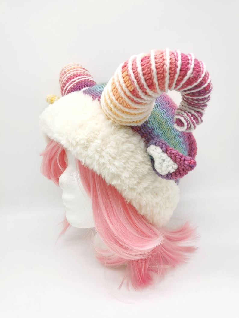 Puede incluir: Un gorro de crochet rosa y blanco con un cuerno grande, rizado y de colores arco&iacute;ris. El gorro est&aacute; hecho con un ribete blanco esponjoso y se lleva sobre una peluca rosa.