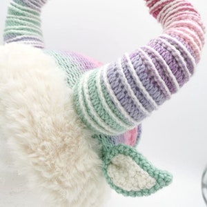 May include: A whimsical, handmade goat horn headpiece. The horns are striped with shades of pink, purple, and green. The base is a soft, white faux fur, with matching green and white crocheted ears. A unique costume accessory.
