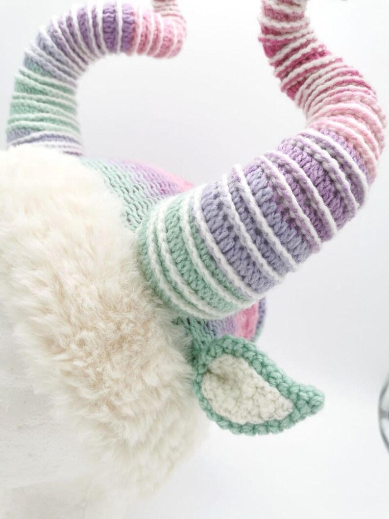 May include: A whimsical, handmade goat horn headpiece. The horns are striped with shades of pink, purple, and green. The base is a soft, white faux fur, with matching green and white crocheted ears. A unique costume accessory.