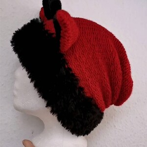 Puede incluir: Un gorro de punto rojo y negro con ribete de piel sint&eacute;tica. El gorro tiene un dise&ntilde;o de punto texturizado y presenta dos peque&ntilde;os cuernos negros en la parte superior. El gorro se exhibe en una cabeza de maniqu&iacute; blanca.