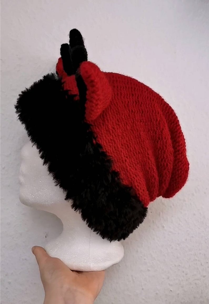 Puede incluir: Un gorro de punto rojo y negro con ribete de piel sint&eacute;tica. El gorro tiene un dise&ntilde;o de punto texturizado y presenta dos peque&ntilde;os cuernos negros en la parte superior. El gorro se exhibe en una cabeza de maniqu&iacute; blanca.