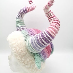 May include: A whimsical, handmade hat featuring two large, spiraling horns in shades of pink, purple, and green. The horns are attached to a fluffy, white faux fur base, creating a unique and fantastical accessory.