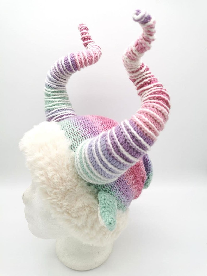 May include: A whimsical, handmade hat featuring two large, spiraling horns in shades of pink, purple, and green. The horns are attached to a fluffy, white faux fur base, creating a unique and fantastical accessory.