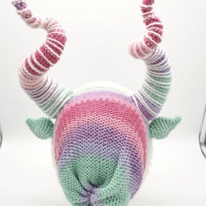 May include: A whimsical knit hat with large, spiraling horns in shades of pink, purple, and white. The hat features a gradient design and small green ears. The horns are approximately 12 inches long, adding a playful touch to the headwear.