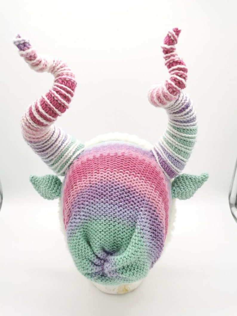 May include: A whimsical knit hat with large, spiraling horns in shades of pink, purple, and white. The hat features a gradient design and small green ears. The horns are approximately 12 inches long, adding a playful touch to the headwear.