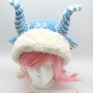 May include: A blue and white knitted hat with a white fur trim and two long, blue and white striped horns. The hat is on a white mannequin head with pink hair.