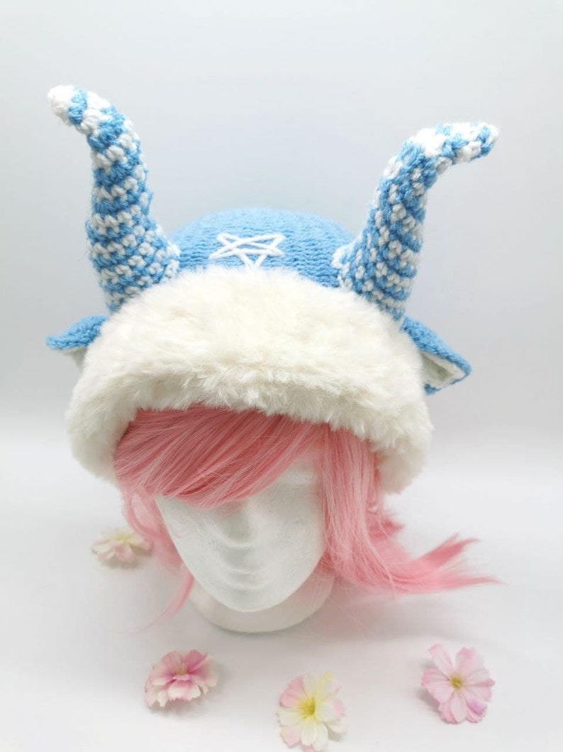 May include: A blue and white knitted hat with a white fur trim and two long, blue and white striped horns. The hat is on a white mannequin head with pink hair.