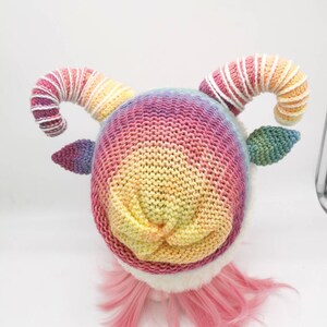 Puede incluir: Un gorro de punto arco&iacute;ris con rayas blancas y rosas en los cuernos. El gorro est&aacute; en una peluca rosa.